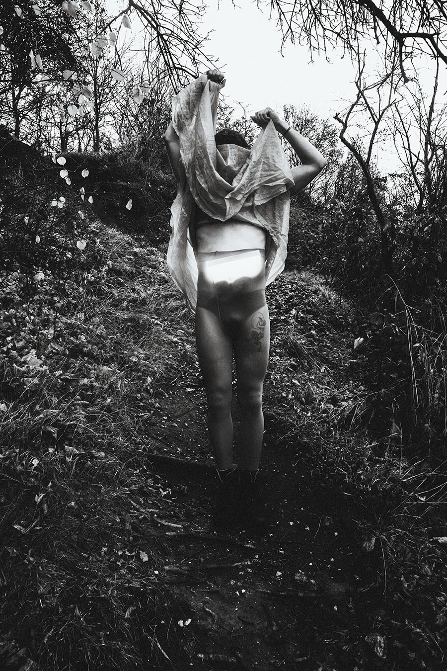 Schwarzweiß-Fotografie einer Person auf einem Waldpfad, die ein Tuch über den Kopf hebt, konzeptuelle Fine-Art-Fotografie.