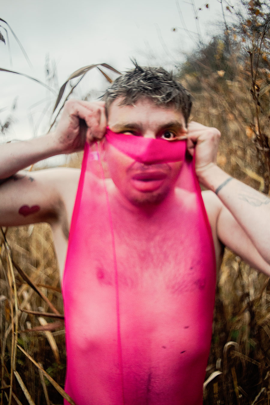 Eine Person steht inmitten hoher Gräser und zieht sich ein enges, pinkes, transparentes Stoffstück über Gesicht und Oberkörper.