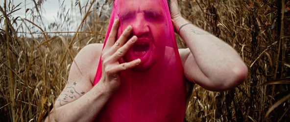 Der Mensch im Bild scheint sich den pinken Stoff aus dem Gesicht zu reißen oder sich darin zu verlieren. Das Umfeld aus dürren Pflanzen verstärkt das Gefühl von Isolation und roher Körperlichkeit. Ein Bild über Enge, Identität, Scham, Wiederstand — oder Transformation.