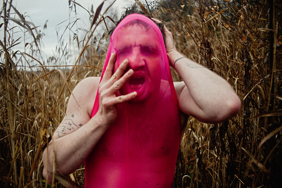 Der Mensch im Bild scheint sich den pinken Stoff aus dem Gesicht zu reißen oder sich darin zu verlieren. Das Umfeld aus dürren Pflanzen verstärkt das Gefühl von Isolation und roher Körperlichkeit. Ein Bild über Enge, Identität, Scham, Wiederstand — oder Transformation.