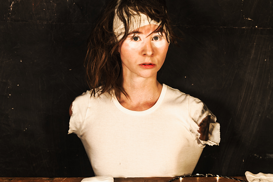 Porträt einer Frau vor dunklem Hintergrund, die mit ernstem, direktem Blick in die Kamera schaut. Sie trägt ein weißes, an den Schultern zerissenes T-Shirt und ein weißes Stirnband; ihre Haare wirken zerzaust. Die Arme sind nicht sichtbar, als wären sie im Stoff verborgen. Vor ihr steht ein Tisch, auf dem ein silbernes Tablett und ein weißes Tuch liegen