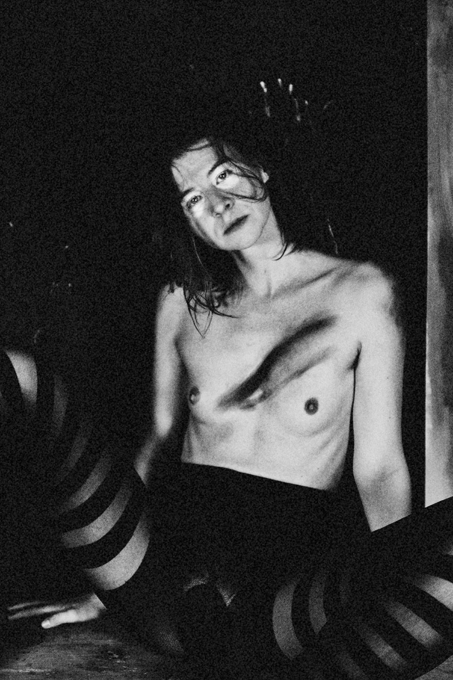 Schwarz-weiß-Fotografie einer sitzenden Person mit nacktem Oberkörper in dunkler Umgebung. Die Person trägt gestreifte Strumpfhosen und hat den Kopf leicht zur Seite geneigt. Das Gesicht wirkt nachdenklich, die Haare fallen teilweise ins Gesicht. Auf der Brust sind dunkle, malerisch wirkende Spuren zu sehen. Die Szene ist kontrastreich und atmosphärisch dunkel.