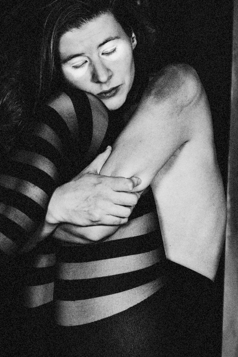 Schwarz-weiß-Porträt einer Person, die mit geschlossenen Augen ihren eigenen Körper umarmt. Sie trägt eine eng anliegende, gestreifte Strumpfhose; Arme und Beine sind ineinander verschlungen. Die Haltung wirkt schützend und introspektiv, das Licht betont Gesicht und Hände vor dunklem Hintergrund.