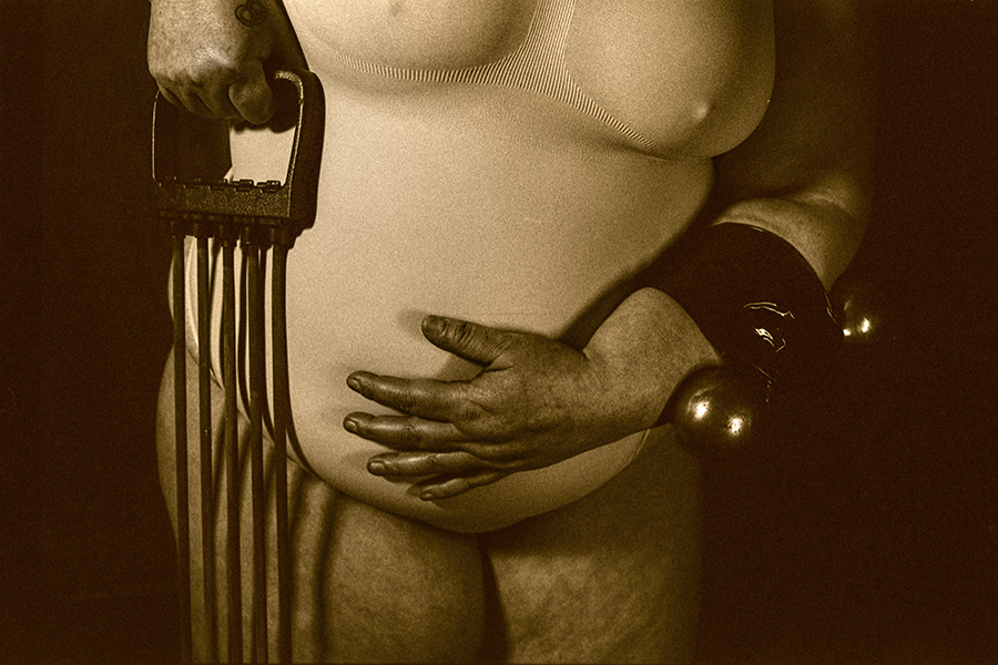 Teilakt einer kurvigen Person in heller Unterwäsche, eine Hand auf dem Bauch, die andere hält ein Trainingsgerät mit Gummibändern; am Arm befestigte Hantel; analoge Schwarzweißaufnahme.