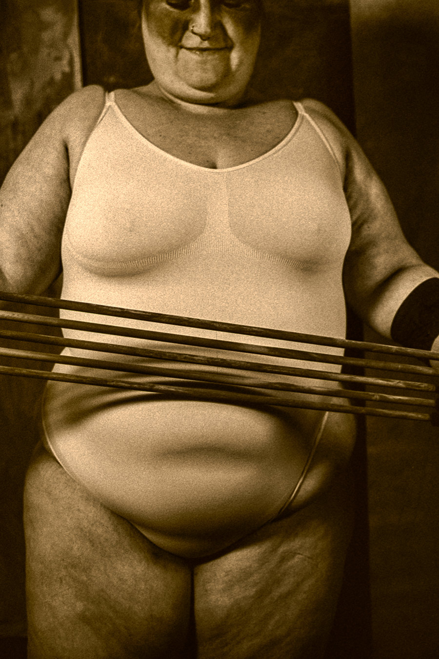 Teilakt einer kurvigen Person in heller Unterwäsche, die mehrere gespannte Stäbe oder Bänder auf Bauchhöhe horizontal hält; analoge Schwarzweißaufnahme mit starkem Kontrast.