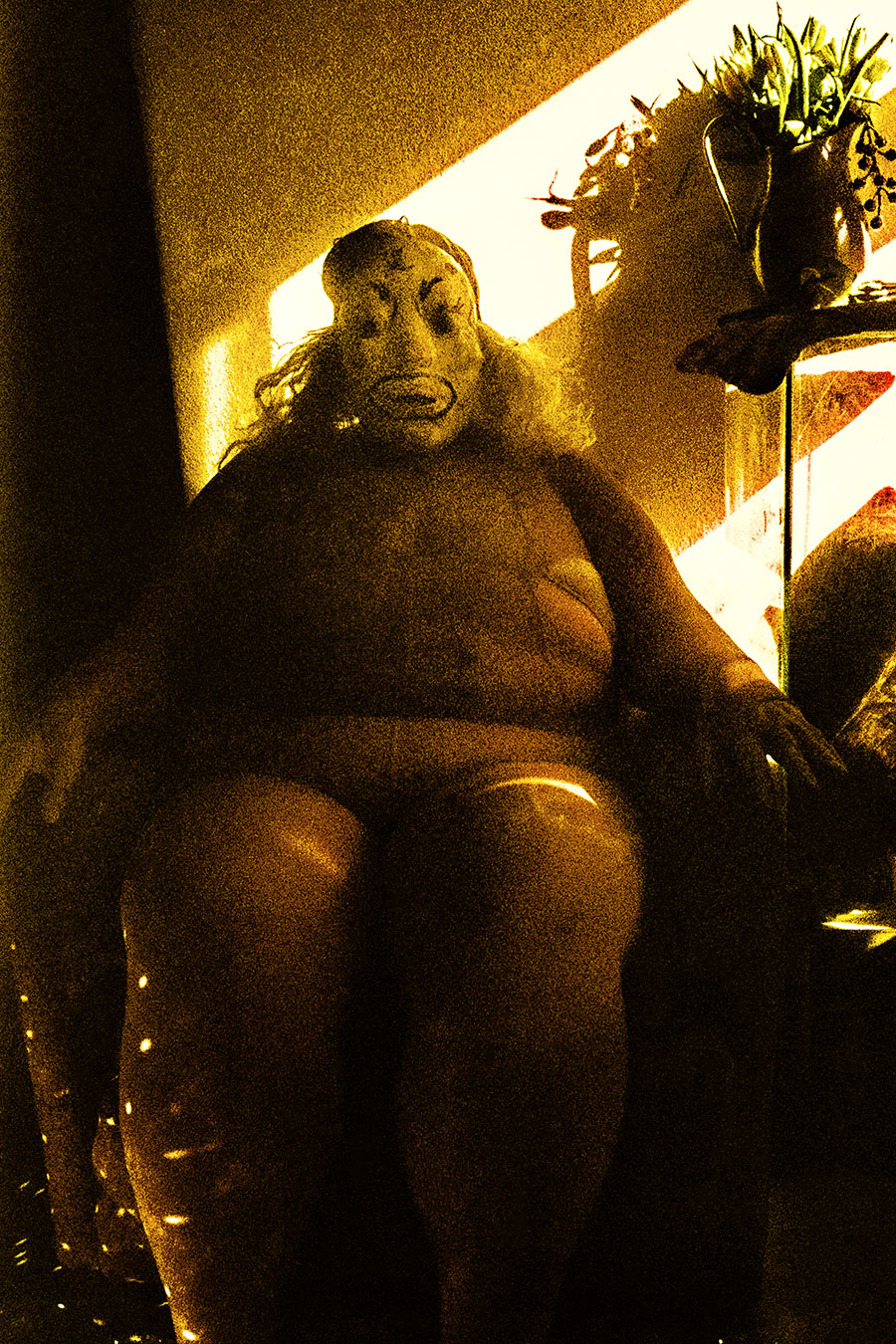Analoges Foto in tiefem Gelb-Schwarz. Eine Person sitzt in einem Sessel, trägt eine Maske mit grotesk-traurigem Ausdruck. Lichterketten am Boden, Pflanze auf einem Regal rechts. Starkes Korn, kaum Details.