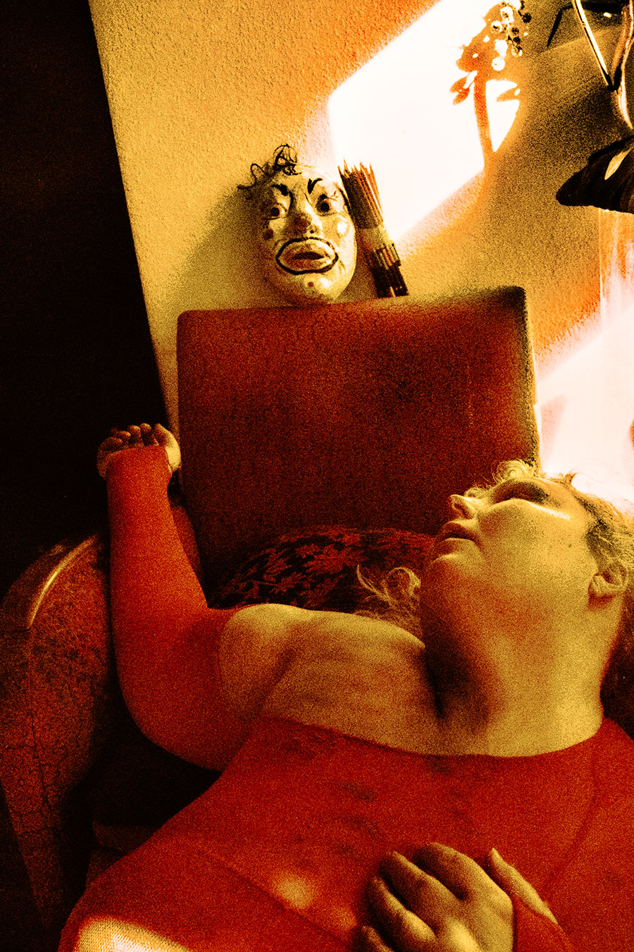 Analoges Foto in Rot-Orange. Eine Person in rotem Top liegt in einem Sessel, Kopf zurückgeworfen, Hals freigelegt. Im Hintergrund hängt eine weiß-bemalte Clownmaske an der Wand, daneben ein Bündel Bleistifte. Oben rechts ein abstrakter oranger Farbfleck.