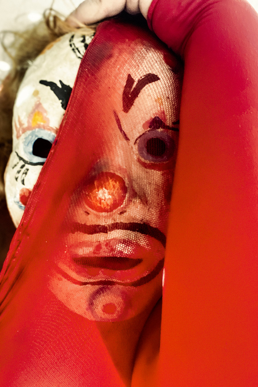 Eine bemalte Clownmaske wird durch roten Stoff hindurch sichtbar — der Stoff liegt transparent über dem Gesicht. Dahinter eine zweite Maske. Die Hand hält beides. Dominante Rottöne, weißer Hintergrund.