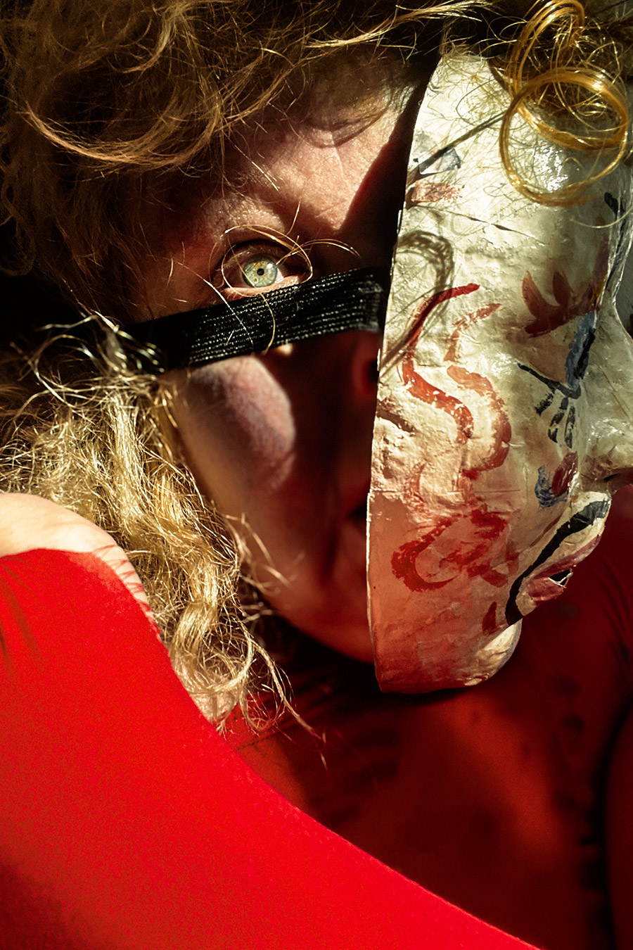 Extremes Nahbild. Ein Auge schaut hinter einer bemalten Maske hervor, die halb das Gesicht verdeckt. Ein schwarzes Band liegt quer über Mund und Nase. Lockiges blondes Haar, rotes Oberteil, hartes Seitenlicht.