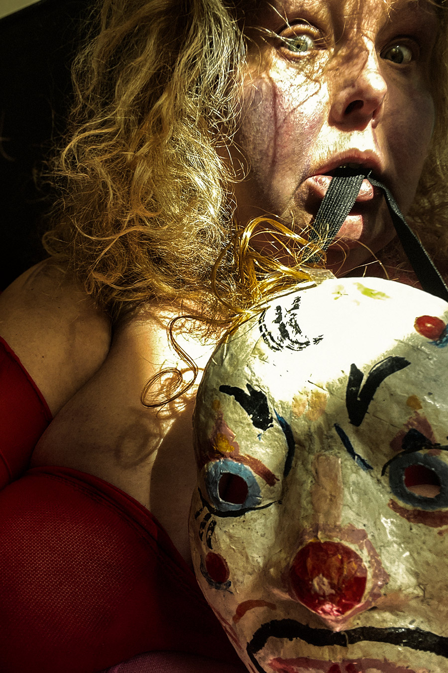 Nahaufnahme. Eine Person hält eine bemalte Clownmaske vor ihre Brust, ein schwarzes Band im Mund. Blondes, zerzaustes Haar mit goldenen Fäden durchsetzt. Weit aufgerissene Augen, leicht geöffneter Mund. Rotes Oberteil, dramatisches Seitenlicht.