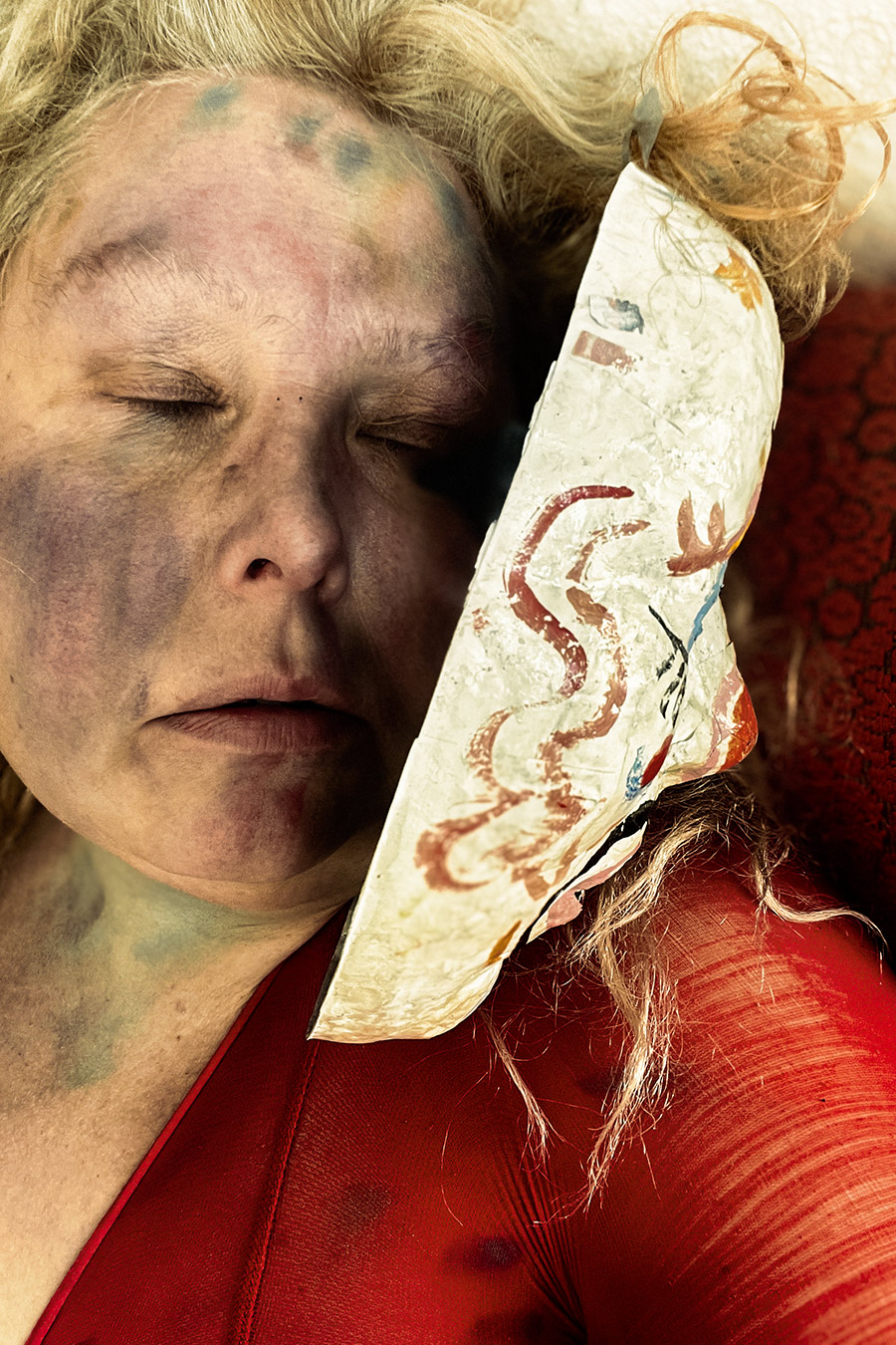 Nahaufnahme eines Gesichts mit geschlossenen Augen und dunklen Farbflecken auf der Haut. Neben dem Gesicht die Innenseite einer bemalten Clownmaske, sichtbar aus dem Blickwinkel der Trägerin. Blondes Haar, rotes Oberteil.