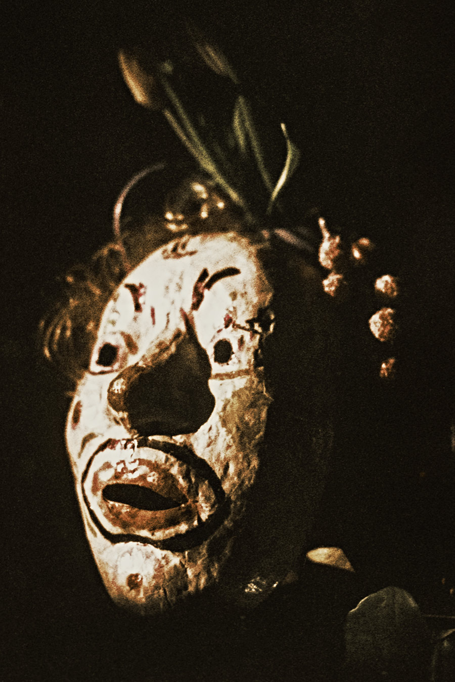 Stark gekörntes Foto vor schwarzem Hintergrund. Eine weiß-bemalte, handgefertigte Clownmaske mit offenem Mund und geritzten Linien, aus dem Kopf Pflanzentriebe. Rechts eine Hand mit ausgestreckten Fingern. Warme, dunkle Töne.