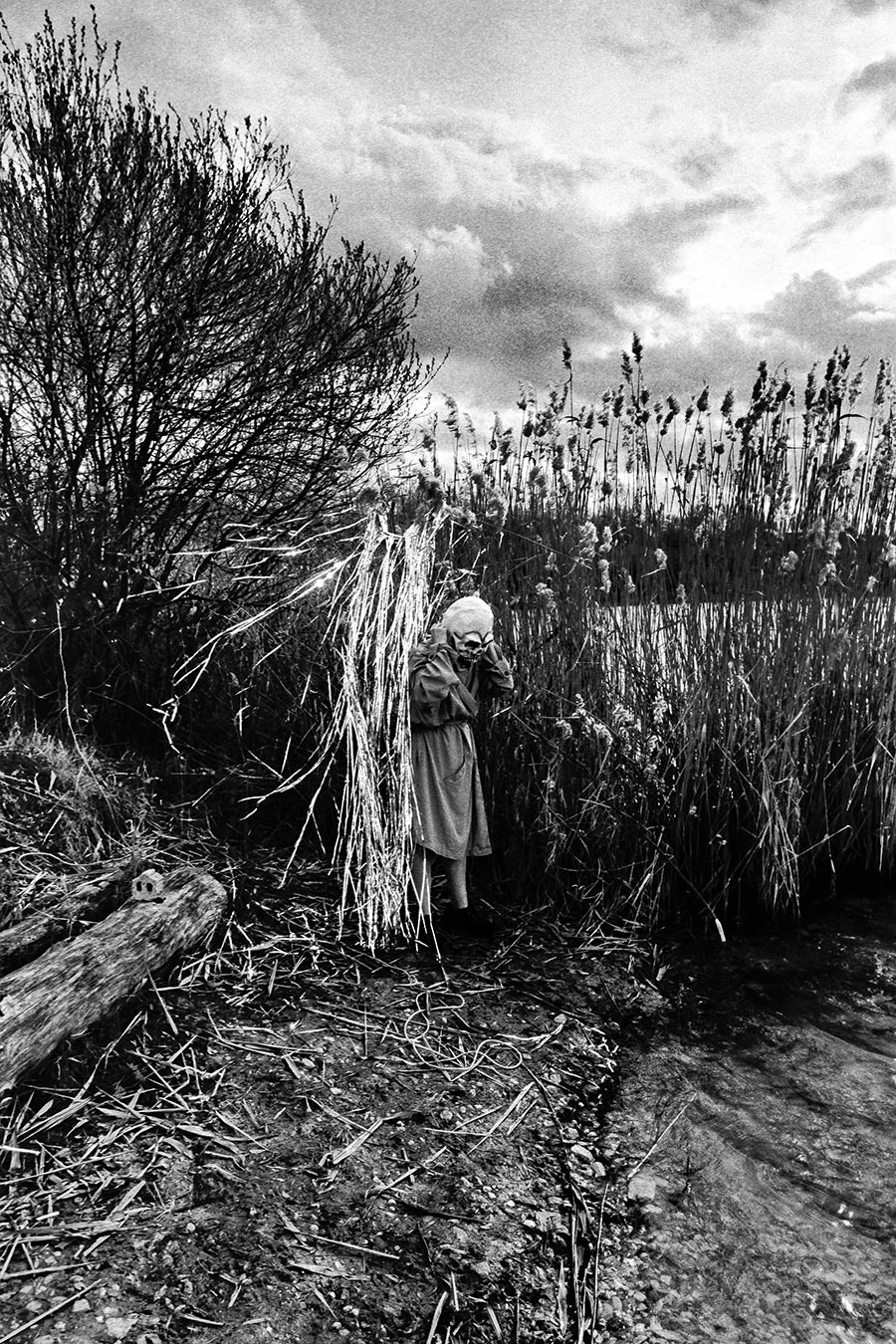 Maskierte Gestalt mit Totenmaske steht zusammengekauert im Schilf am Wasser, Silberlametta, analoges Schwarz-Weiß-Foto auf Kodak T-Max 100, Serie „El Pasaje"