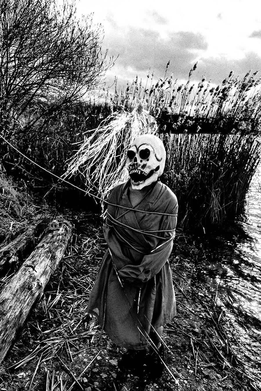 Maskierte Gestalt mit Seil gefesselt und Silberlametta am Kopf, steht im Schilf am Wasser, bewölkter Himmel, analoges Schwarz-Weiß-Foto auf Kodak T-Max 100, Serie „El Pasaje"