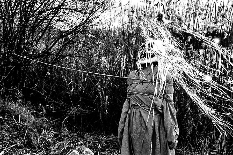 Maskierte Gestalt mit Seil gefesselt, überwältigt von Silberlametta-Bündel, Schilf und Seil im Vordergrund, analoges Schwarz-Weiß-Foto auf Kodak T-Max 100, Serie „El Pasaje"