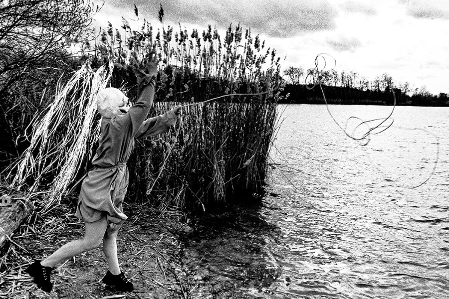 Maskierte Gestalt wirft Seil ins Wasser, in Bewegung, Schilf und See im Hintergrund, analoges Schwarz-Weiß-Foto auf Kodak T-Max 100, Serie „El Pasaje"