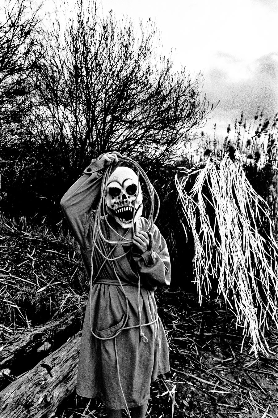 Maskierte Gestalt hält Seil in Händen, steht vor Silberlametta-Bündel und kahlen Ästen, analoges Schwarz-Weiß-Foto auf Kodak T-Max 100, Serie „El Pasaje"