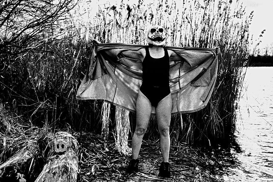 Frau mit Totenmaske breitet Umhang aus, steht im Badekostüm am Seeufer, Schilf hinter ihr, analoges Schwarz-Weiß-Foto auf Kodak T-Max 100, Serie „El Pasaje"