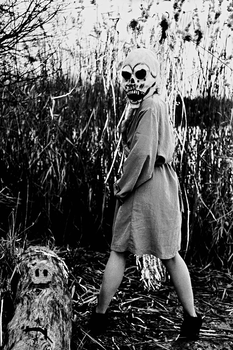 Maskierte Gestalt mit Totenmaske steht seitlich im Schilf, Silberlametta hängt herab, Treibholz im Vordergrund, analoges Schwarz-Weiß-Foto auf Kodak T-Max 100, Serie „El Pasaje"