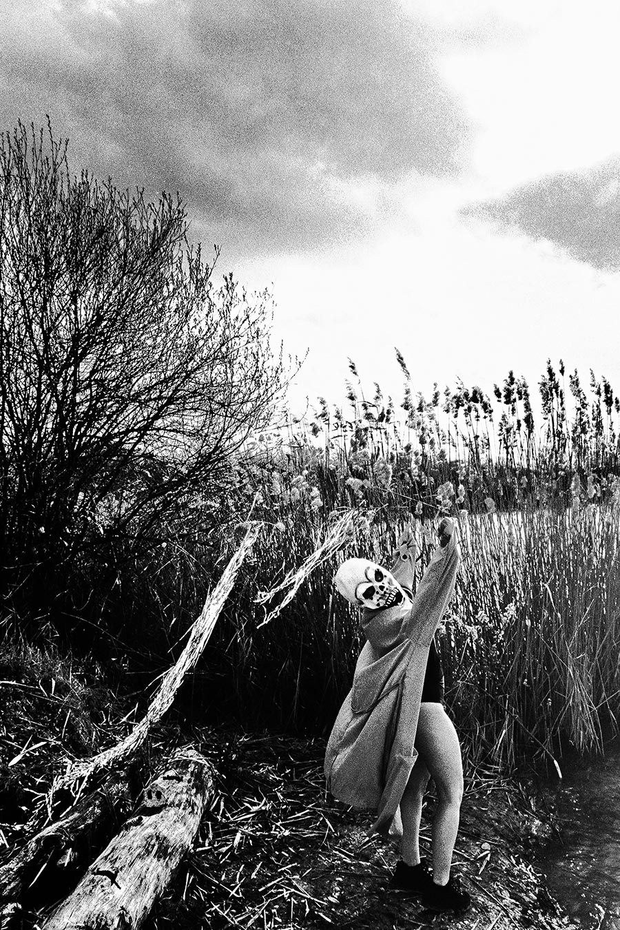 Maskierte Gestalt kauert am Ufer und greift in das Wasser, Schilf und Treibholz, analoges Schwarz-Weiß-Foto auf Kodak T-Max 100, Serie „El Pasaje"