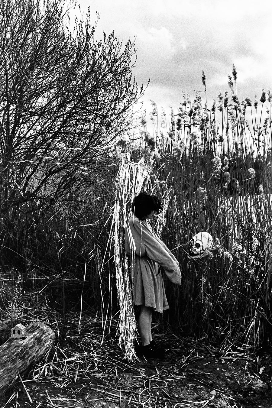 Frau ohne Maske steht im Schilf, hält Schädel in der Hand, daneben Silberlametta-Bündel, analoges Schwarz-Weiß-Foto auf Kodak T-Max 100, Serie „El Pasaje"