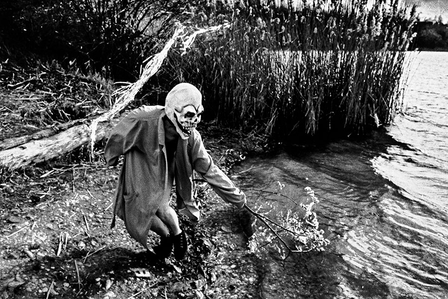 Maskierte Gestalt hält blühenden Zweig zur Totenmaske, steht am Seeufer mit Schilf, analoges Schwarz-Weiß-Foto auf Kodak T-Max 100, Serie „El Pasaje"
