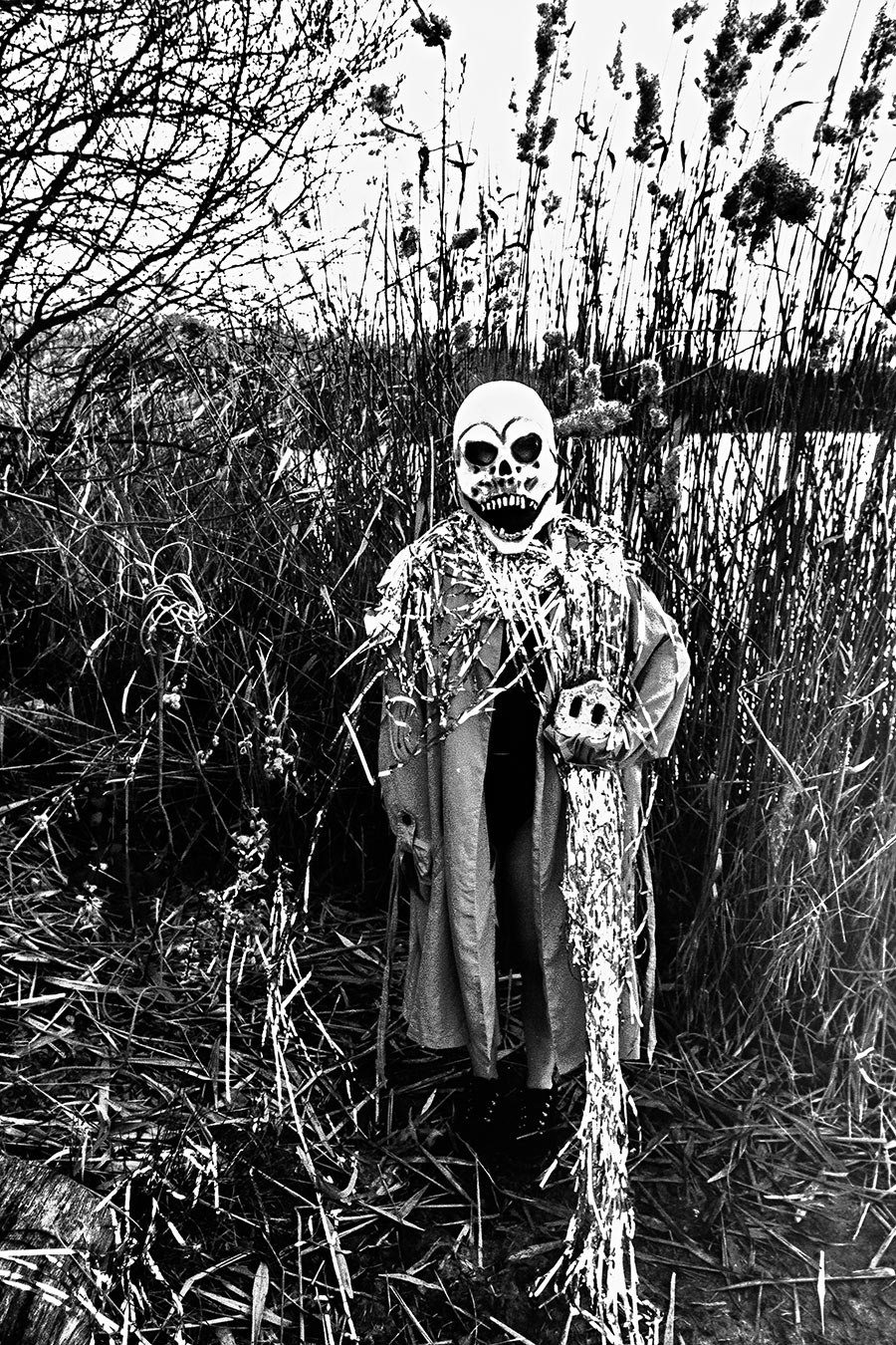 Maskierte Gestalt steht frontal im Schilf, Silberlametta am Hals, hält Ziegelstein, analoges Schwarz-Weiß-Foto auf Kodak T-Max 100, Serie „El Pasaje"