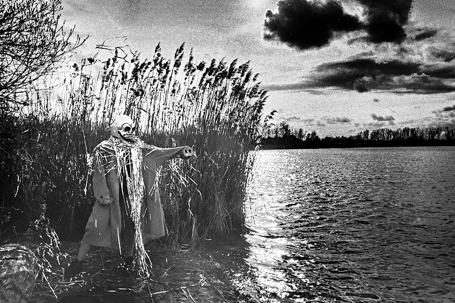 Maskierte Gestalt am Seeufer streckt Arm über das Wasser aus, Silberlametta, dramatischer Wolkenhimmel, analoges Schwarz-Weiß-Foto auf Kodak T-Max 100, Serie „El Pasaje"