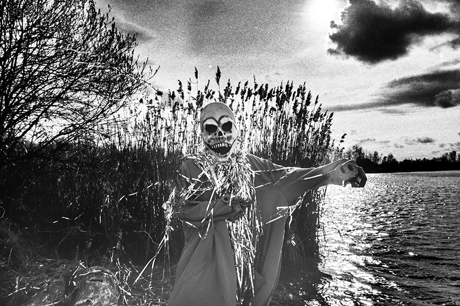 Maskierte Gestalt mit Silberlametta steht frontal am Seeufer, Schilf, Wolkenhimmel, starkes Filmkorn, analoges Schwarz-Weiß-Foto auf Kodak T-Max 100, Serie „El Pasaje"