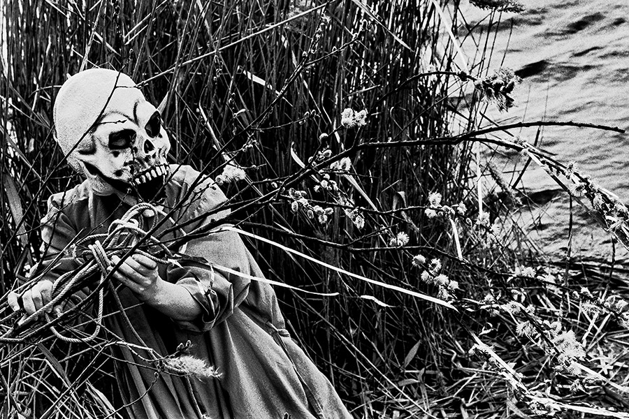 Maskierte Gestalt mit Seil hält Weidenzweig, sitzt im Schilf am Wasser, Nahaufnahme, analoges Schwarz-Weiß-Foto auf Kodak T-Max 100, Serie „El Pasaje"