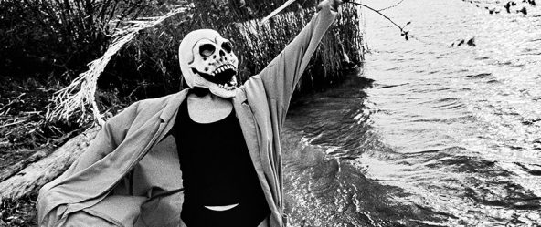 Maskierte Gestalt hält blühenden Ast mit Kätzchen, steht vor Treibholz und Schilf am Wasser, analoges Schwarz-Weiß-Foto auf Kodak T-Max 100, Serie „El Pasaje"