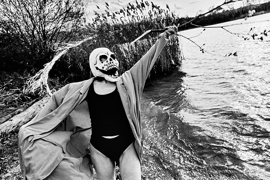 Maskierte Gestalt hält blühenden Ast mit Kätzchen, steht vor Treibholz und Schilf am Wasser, analoges Schwarz-Weiß-Foto auf Kodak T-Max 100, Serie „El Pasaje"