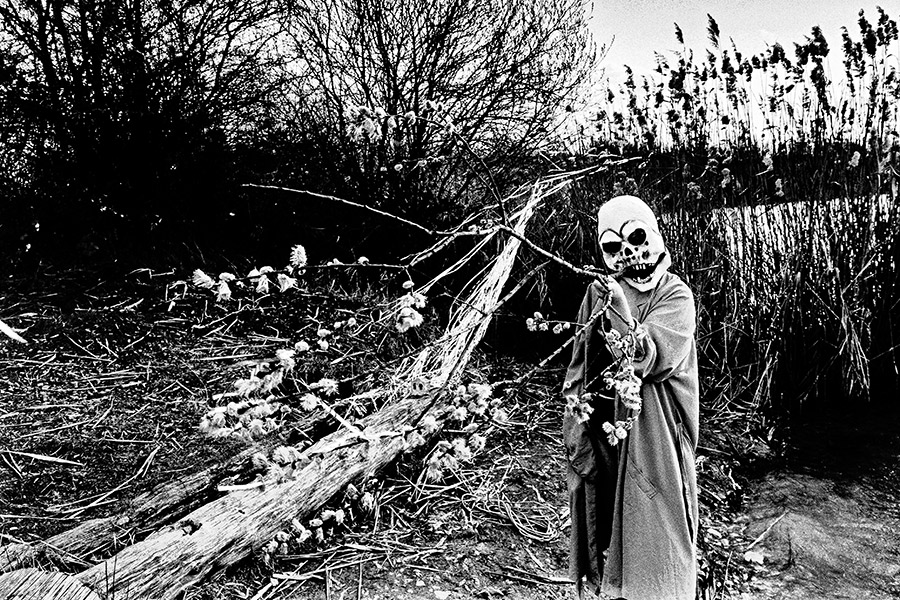 Maskierte Gestalt im Badekostüm greift nach kahlem Ast über dem Wasser, Umhang offen, analoges Schwarz-Weiß-Foto auf Kodak T-Max 100, Serie „El Pasaje"