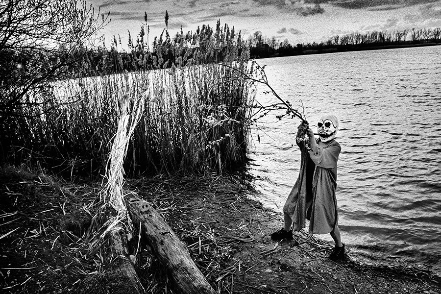 Maskierte Gestalt am Seeufer greift nach Ast, weite Landschaft mit Treibholz und Silberlametta-Bündel, analoges Schwarz-Weiß-Foto auf Kodak T-Max 100, Serie „El Pasaje"