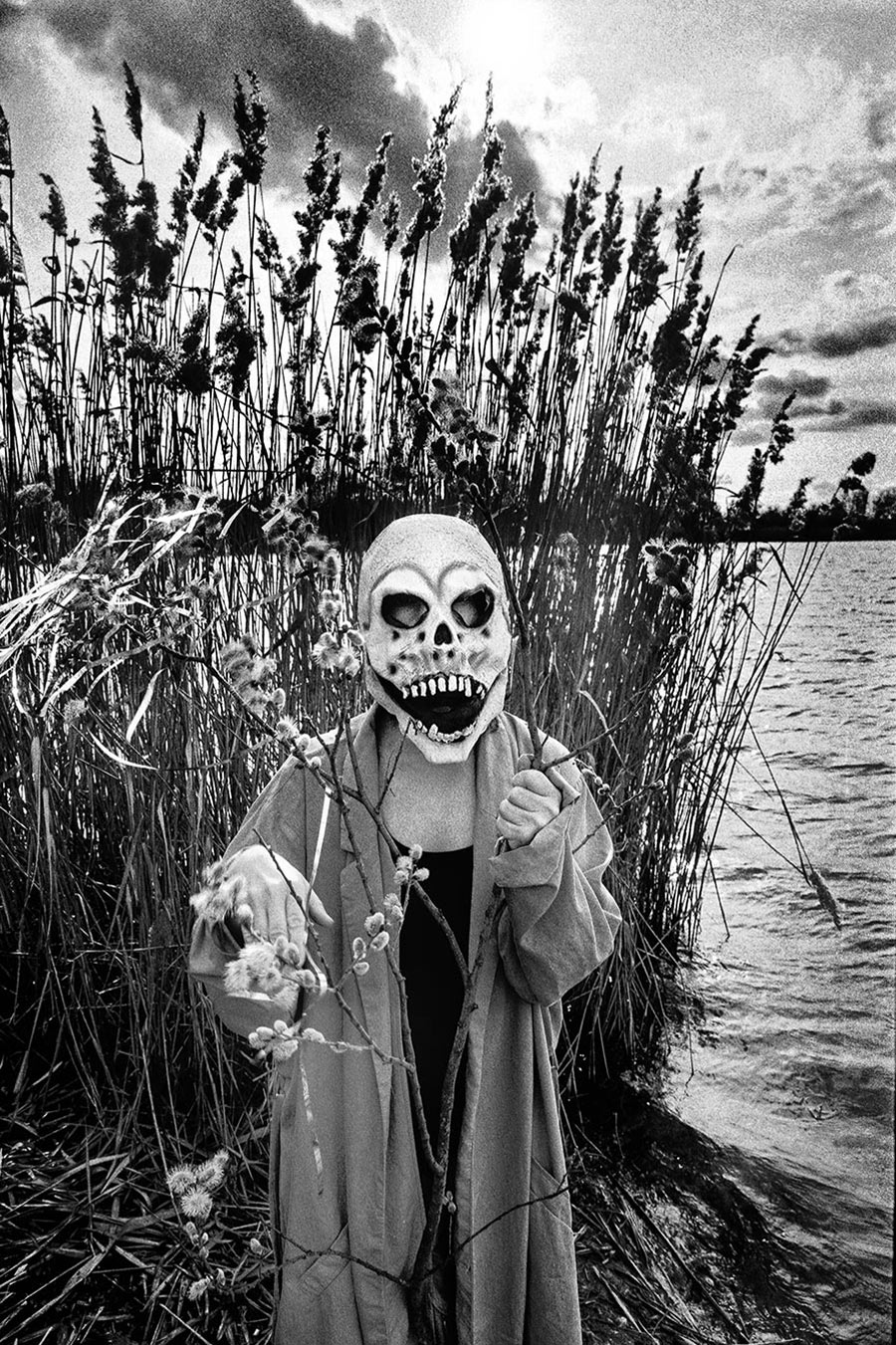 Maskierte Gestalt am Seeufer hält blühenden Weidenzweig hoch, weite Landschaft mit Schilf und Treibholz, analoges Schwarz-Weiß-Foto auf Kodak T-Max 100, Serie „El Pasaje“