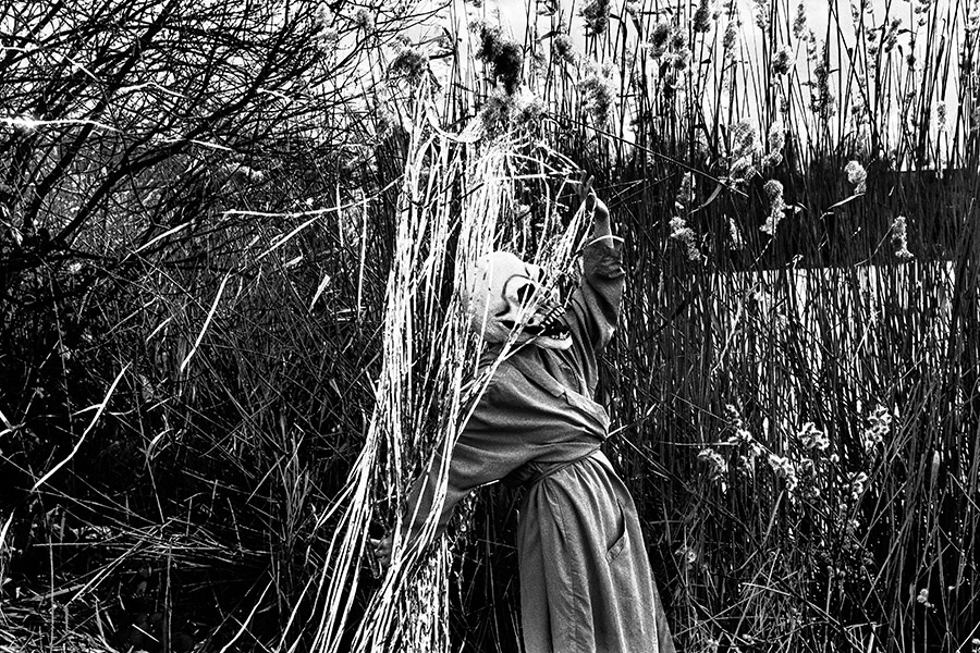 Maskierte Gestalt im Profil, hält Silberlametta-Bündel, Schilf im Hintergrund, analoges Schwarz-Weiß-Foto auf Kodak T-Max 100, Serie „El Pasaje"