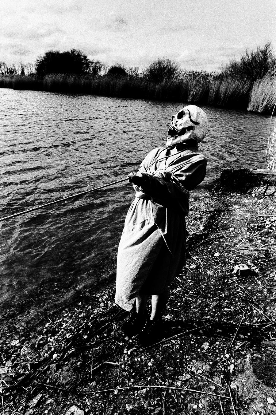 Maskierte Gestalt mit Seil gefesselt steht am Seeufer, Kopf zurückgeworfen, weite Wasserfläche, analoges Schwarz-Weiß-Foto auf Kodak T-Max 100, Serie „El Pasaje"