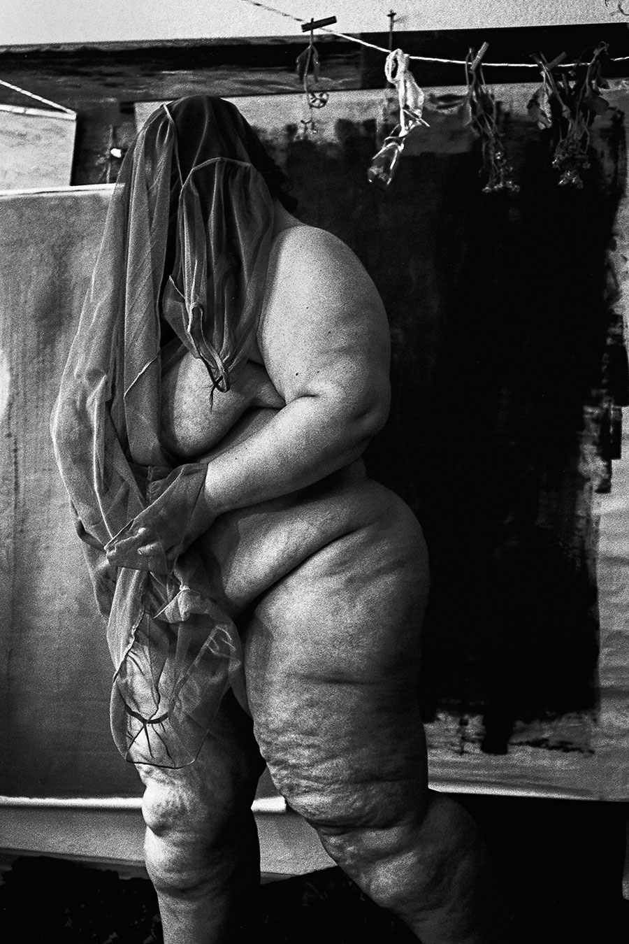 Stehende Frau mit über den Kopf gezogenem Stoff, nackter Körper sichtbar, getrocknete Blumen oben, analoges Schwarz-Weiß-Foto auf Kodak T-Max 100, Serie „Der Ballast"

