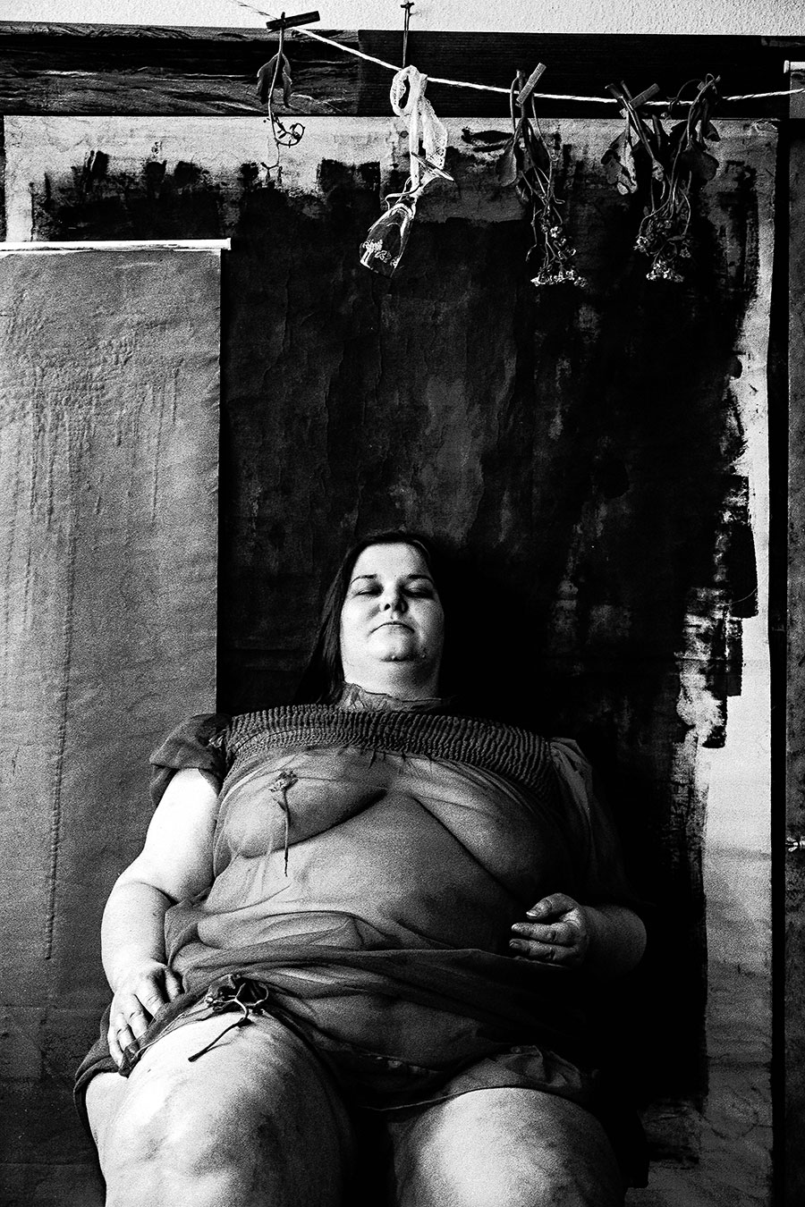 Frau in transparentem Kleid sitzt angelehnt mit geschlossenen Augen, getrocknete Blumen und Objekte hängen über ihr, analoges Schwarz-Weiß-Foto auf Kodak T-Max 100, Serie „Der Ballast"
