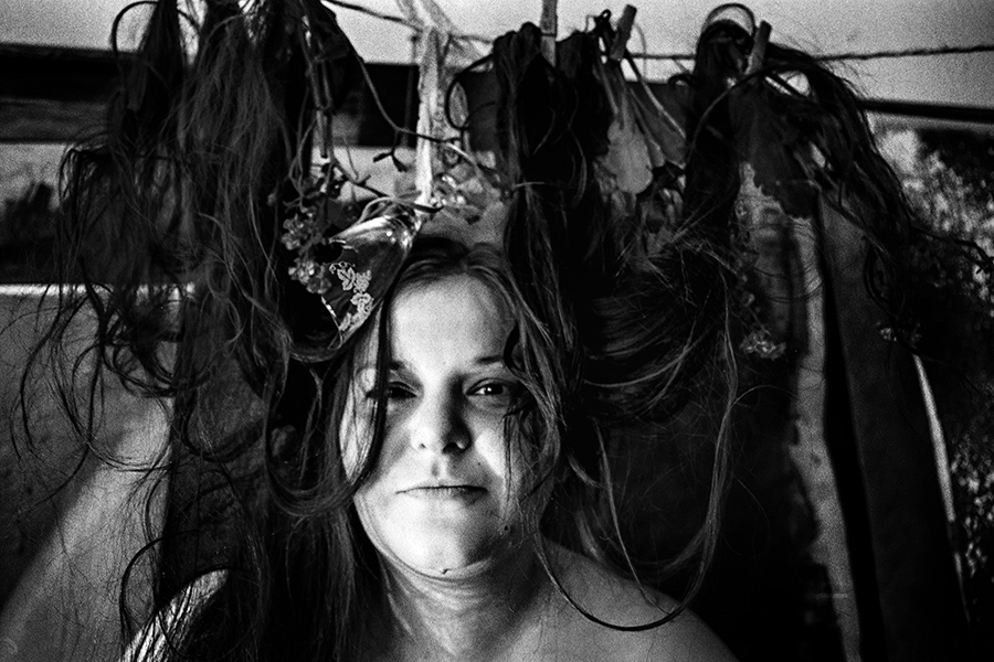 Frau schaut direkt in die Kamera, Haare und Objekte hängen an der Leine über ihr, Nahaufnahme, analoges Schwarz-Weiß-Foto auf Kodak T-Max 100, Serie „Der Ballast"
