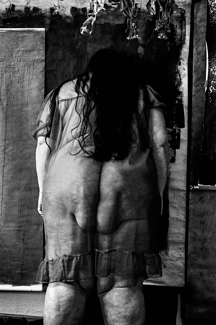 Rückenansicht einer Frau in transparentem Kleid vor bemalten Leinwänden, getrocknete Blumen hängen oben, analoges Schwarz-Weiß-Foto auf Kodak T-Max 100, Serie „Der Ballast"