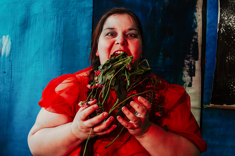 Frau in rotem Kleid mit geflochtenim Zopf beißt in Blumen, blauer Hintergrund, Unschärfe, Serie „Der Ballast"