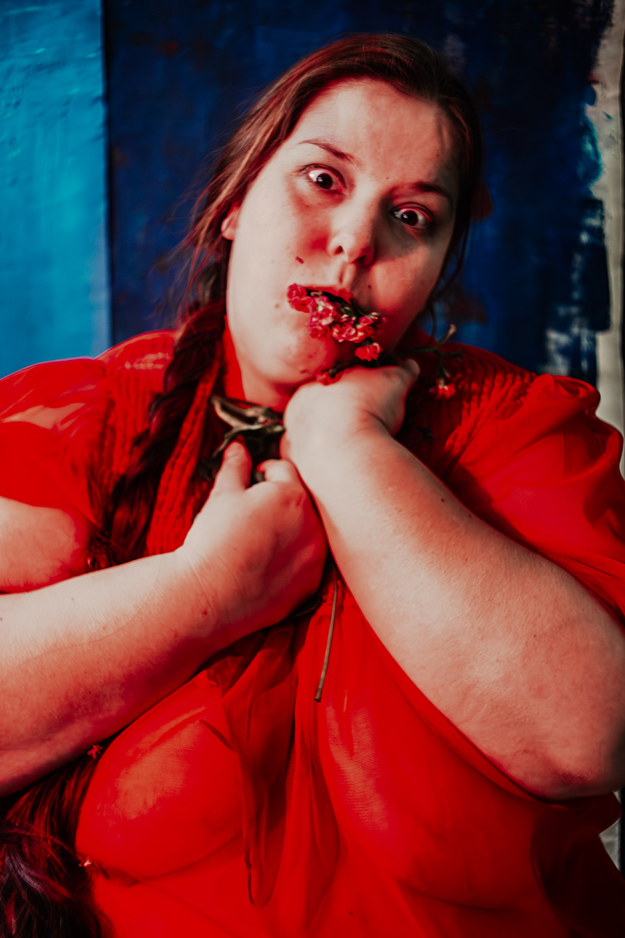 Frau in rotem Kleid mit geflochtenim Zopf beißt in Blumen, blauer Hintergrund, Unschärfe, Serie „Der Ballast"
