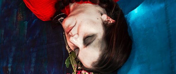 Kopf einer Frau von oben, liegend, mit Blumen am Mund, roter Stoff und blauer Hintergrund, Serie „Der Ballast"