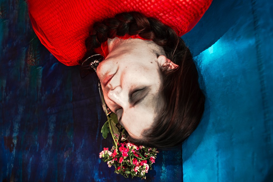 Kopf einer Frau von oben, liegend, mit Blumen am Mund, roter Stoff und blauer Hintergrund, Serie „Der Ballast"