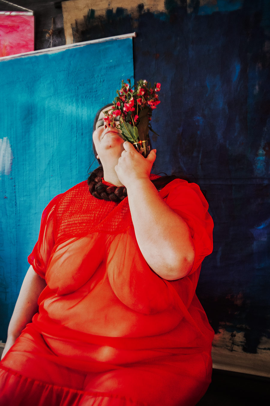 Frau in halb geöffnetem rotem Kleid sitzt mit Blumen am Hals vor blauem Hintergrund, Serie „Der Ballast"