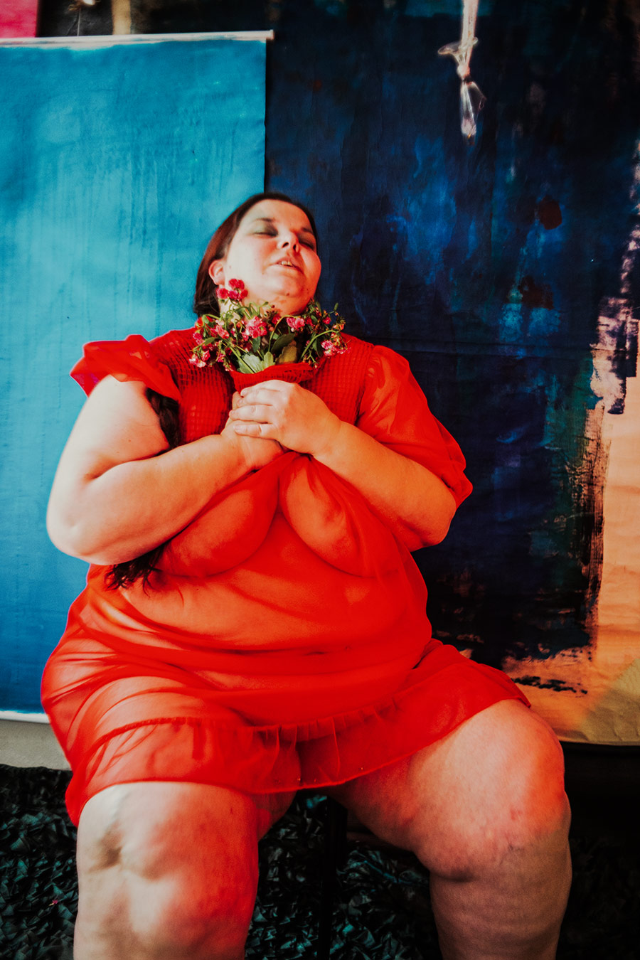 Frau in rotem Kleid mit Zopf hält Blumen ans Gesicht und blickt nach oben, blauer Hintergrund, Serie „Der Ballast"