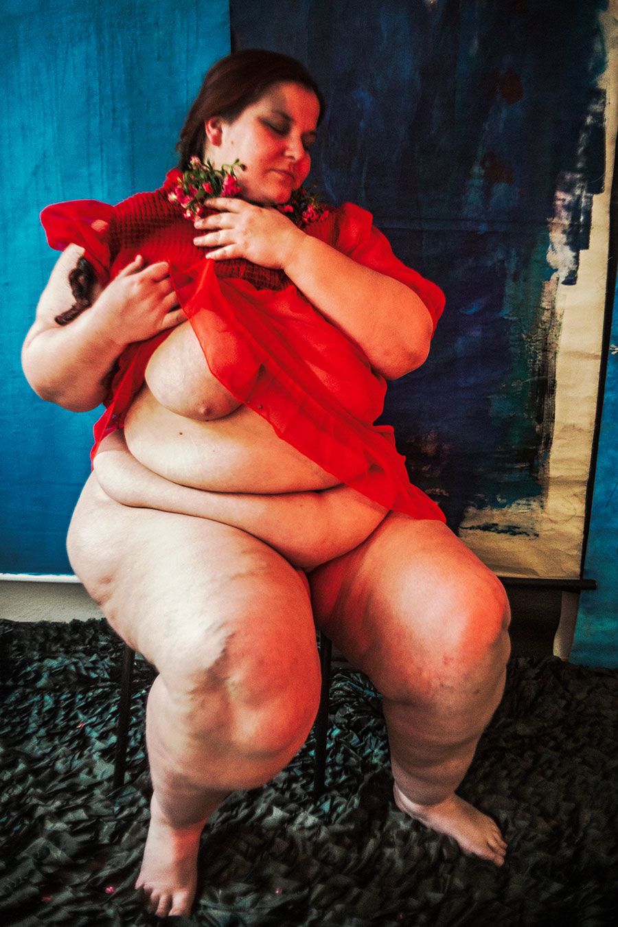Nackte, dicke Frau in rotem Kleid sitzt mit geschlossenen Augen und Blumen am Mund, Zopf seitlich, blauer Hintergrund, Serie „Der Ballast"
