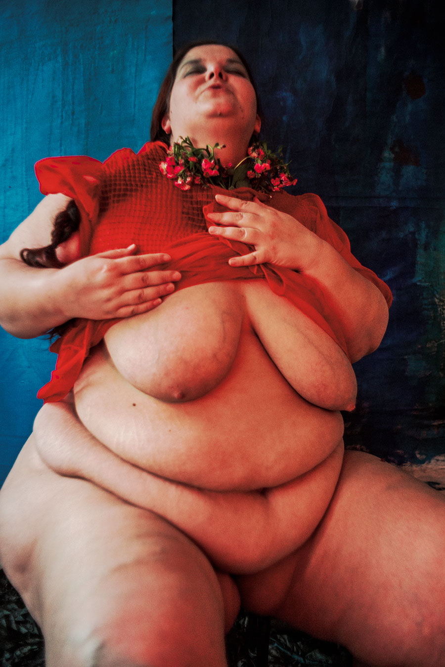 Ganzkörperaufnahme einer dicken Frau in rotem Kleid, die sich gegen eine Wand lehnt und Blumen ans Gesicht hält, blauer Hintergrund, Serie „Der Ballast", ihre großen Brüste sind zu sehen