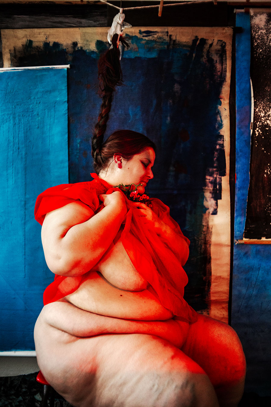 Dicke Frau in geöffnetem rotem Kleid sitzt seitlich mit geflochtenim Zopf und roten Beeren, blauer Hintergrund, Serie „Der Ballast" - ihr Körperfett ist zu sehen